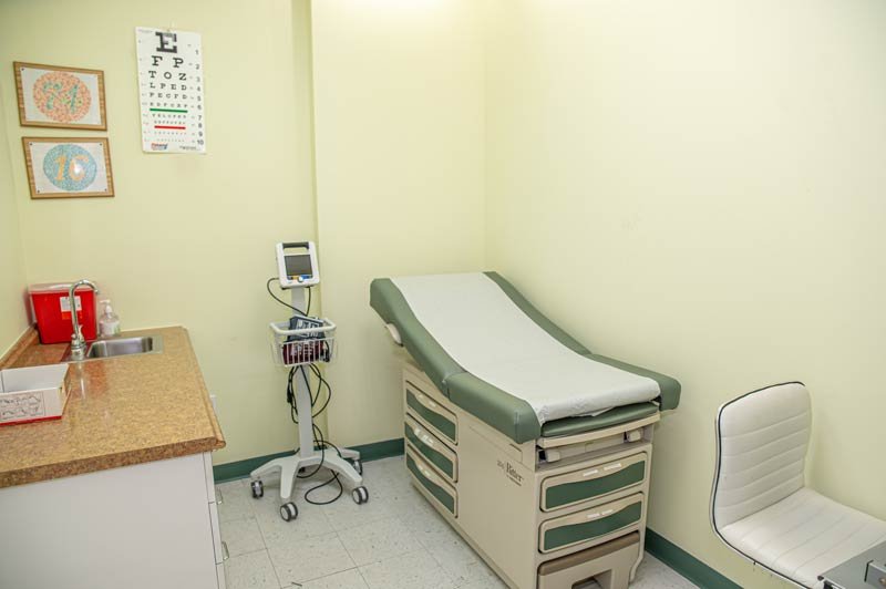 A small medical exam room designed for a DOT Medical Exam, featuring a green and beige exam table, white chair, sink with countertop, medical supplies, wall-mounted eye chart, and digital blood pressure monitor on a stand. DOT Medical Card NYC at Fast CDL Medical