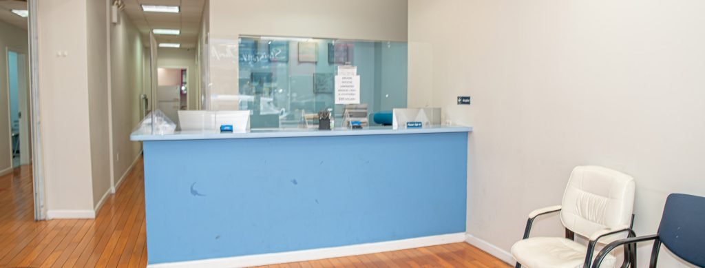 A reception area with a blue counter, clear divider, and contact signs. Two chairs are positioned against a cream-colored wall on a wooden floor. A hallway with doors is visible in the background. DOT Medical Card NYC at Fast CDL Medical