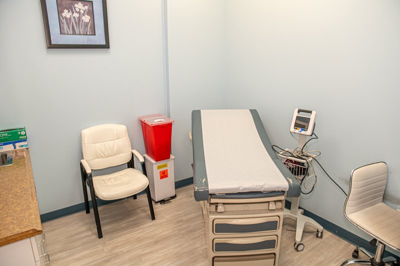 A small medical examination room for DOT Medical Exams with an exam table, two chairs, a medical monitor, a red biohazard waste bin, and a framed picture on a light blue wall. The floor is light wood, and the room appears clean and tidy. DOT Medical Card NYC at Fast CDL Medical